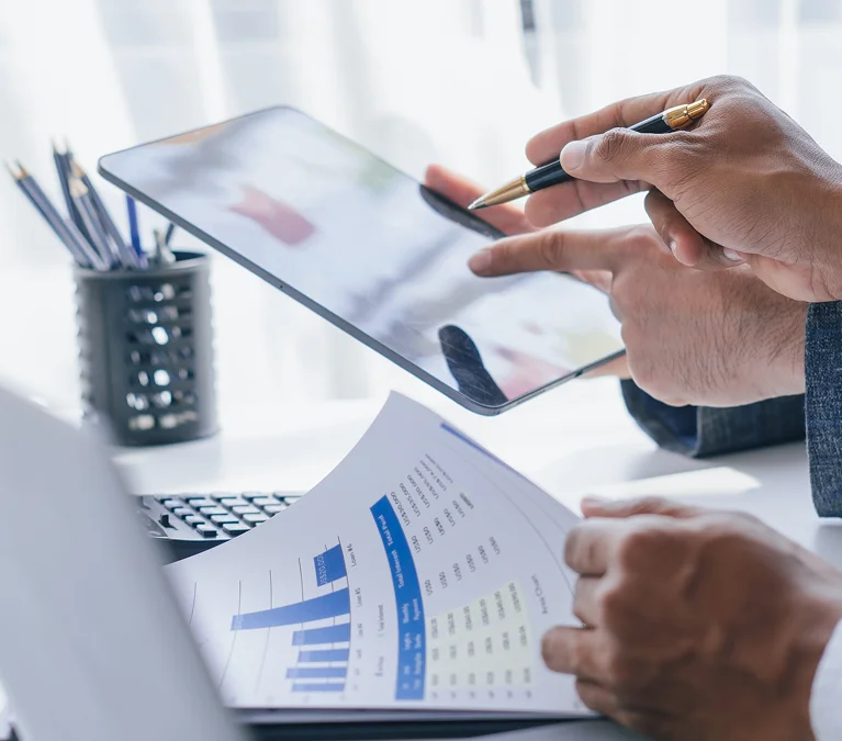 Two people in business attire review charts on a tablet and printed paper at a desk with a laptop and a pencil holder in the background.