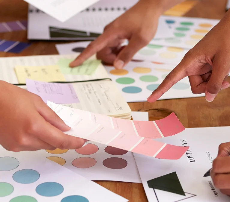 Hands of several people working on a design project, with color swatches, charts, and notes scattered on a table.