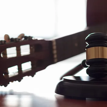 A close-up of a gavel in the foreground, with a blurred guitar neck in the background, symbolizing justice and music.