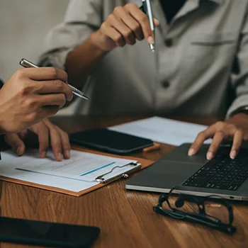 A person’s hand holds a pen above documents on a clipboard, while another hand gestures nearby, suggesting collaboration or discussion.