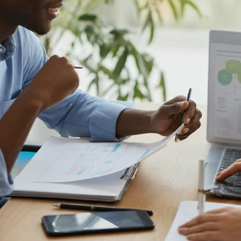 A close-up of two professionals discussing documents at a table, with one using a laptop and the other holding a pen.