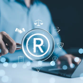 A hand holds a magnifying glass over a glowing registered trademark symbol, surrounded by icons representing business and legal concepts.
